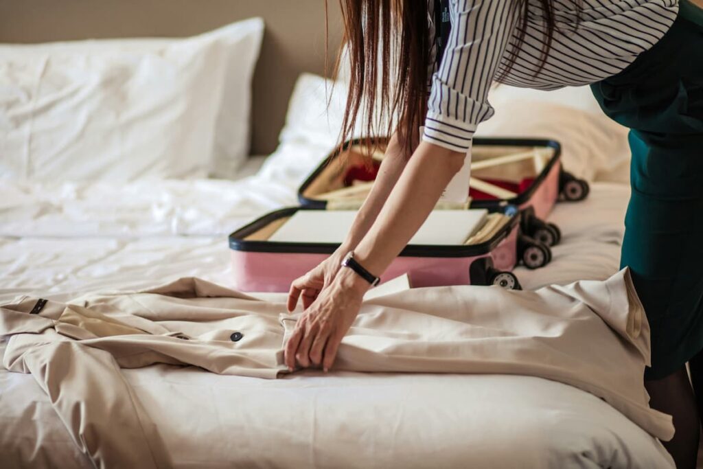 Mulher dobrando um casaco em uma cama, enquanto prepara uma mala de viagem em um ambiente de hotel. A imagem representa planejamento e organização.