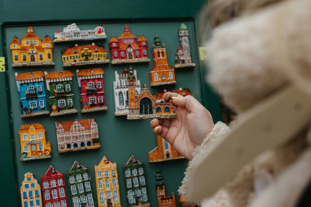 Pessoa segurando um ímã de Gdańsk diante de uma coleção de lembranças da cidade em uma prateleira. A imagem destaca a arquitetura colorida e as popularidades locais.