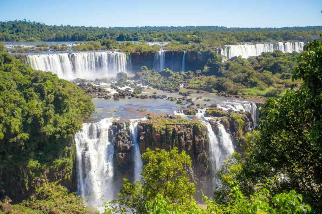 Vista panorâmica das Cataratas do Iguaçu, com várias quedas d'água cercadas por vegetação exuberante em um dia ensolarado.